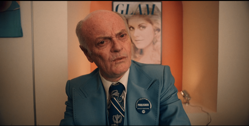 An older white man in a suit looking confused. Behind him is a cover for GLAM magazine with a white woman who seems to be raising one eyebrow.