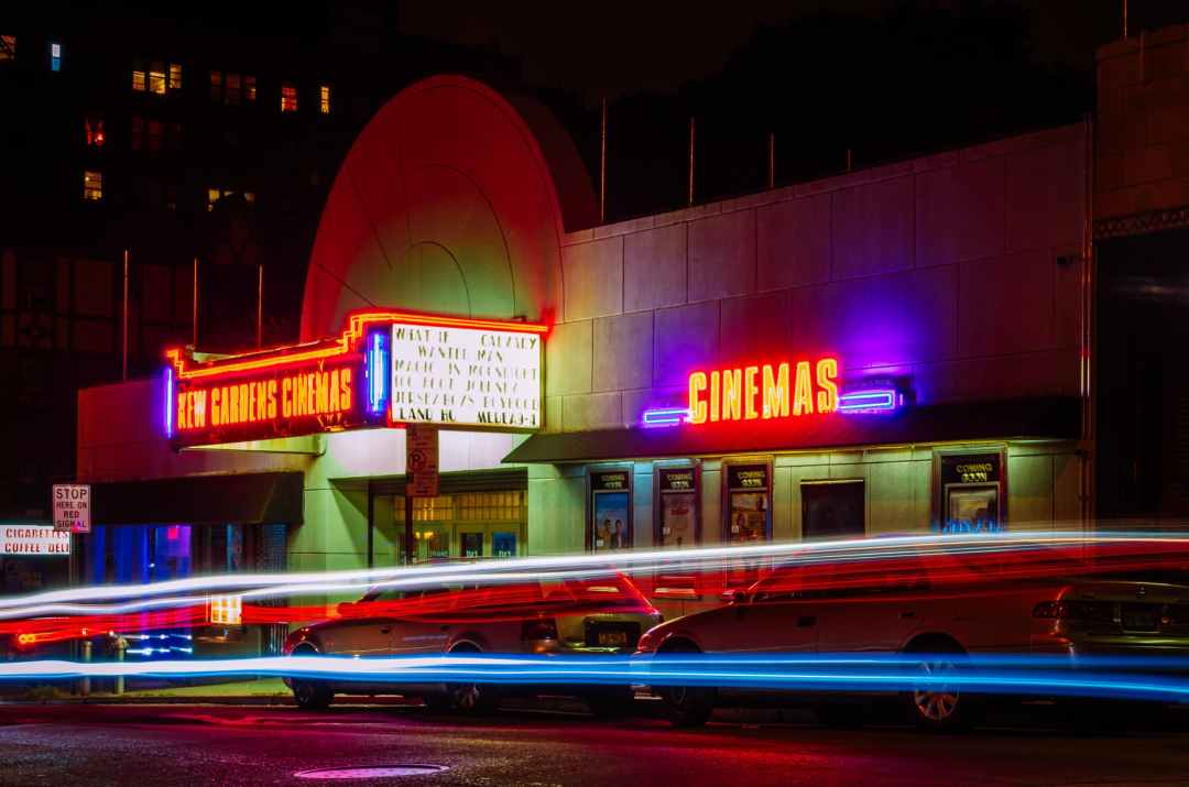 Cinema lit up with neon lights. In front cars are shown in time lapse photography.
