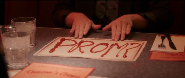 Mandy's hands on the edge of a paper placement on which is written "PROM?" in ketchup as it lays on the table at the diner.